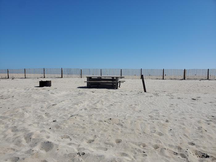 Oceanside site 94 in October.  View of wooden picnic table and black metal fire ring on the sand.  Dune fencing runs along the beach behind the campsite.  Sign post nearby that says 94 on it.Oceanside site 94 in October.