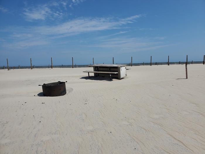 Oceanside site 94 in August.  View of the wooden picnic table and the black metal fire ring on the sand.  Sign post nearby with dune fencing behind the campsite.  Ocean on the horizon.Oceanside site 94 in August.