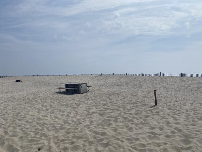 Oceanside site 95 in July 2024.  View of wooden picnic table on the sand.  Signpost nearby that says "95" on it.  Dune fencing runs along the beach front behind the campsite.  Other campsites within the view.Oceanside site 95 - July 2024.