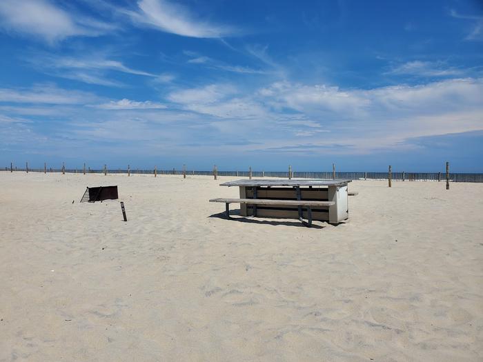 Oceanside site 95 in July 2023.  View of wooden picnic table and black metal fire ring on the sand.  Signpost nearby says "95" on it.  Dune fencing runs along the beach front.  Ocean is on the horizon.Oceanside site 95 - July 2023.