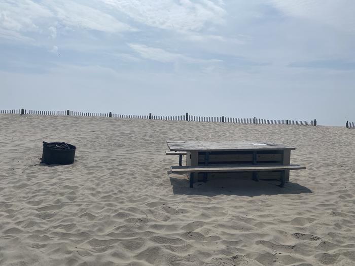 Oceanside site 95 in July 2024.  View of wooden picnic table and black metal fire ring on the sand.  Dune fencing runs along the beach front behind the campsite with a break in the fencing leading to the ocean front to the right of the image.Oceanside site 95 - July 2024.