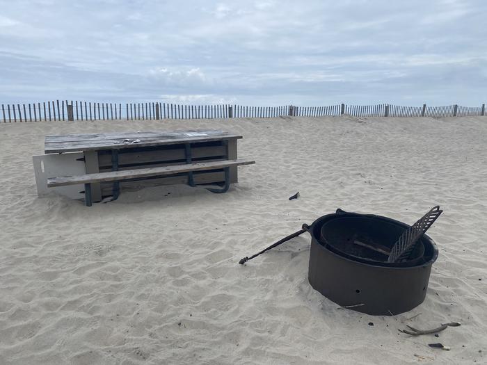 Oceanside site 96 in May.  Close up view of black metal fire ring on the sand.  Wooden picnic table is located behind the fire ring.  Dune fencing runs along the beach front with ocean on the horizon.Oceanside site 96 in May.