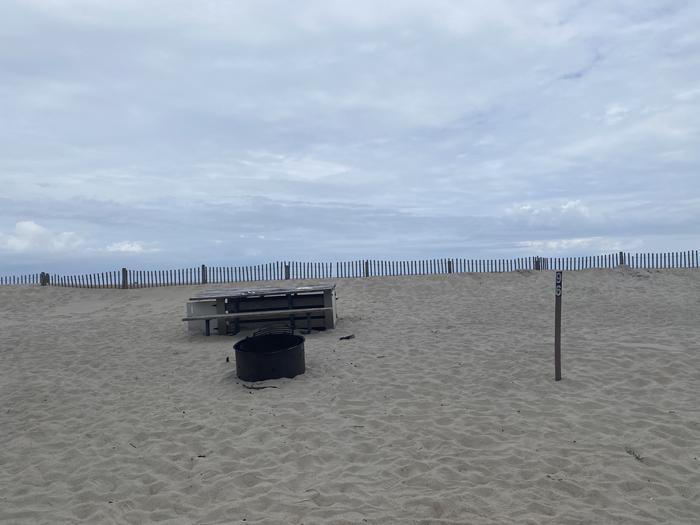 Oceanside site 96 in May.  View of black metal fire ring on the sand.  Wooden picnic table is located behind the fire ring.  Sign post nearby says 96 on it.  Dune fencing runs along the beach behind the campsite.  Oceanside site 96 in May.