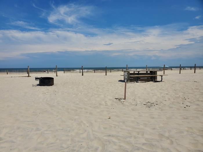 Oceanside site 96 in July.  View of the wooden picnic table and black metal fire ring on the sand.  Sign post nearby says 96 on it.  Dune fencing runs along the back of the campsite.  Ocean on the horizon.Oceanside site 96 in July.