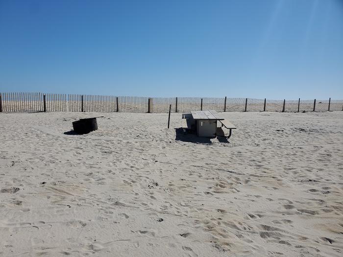 Oceanside site 96 in October.  View of the wooden picnic table and black metal fire ring on the sand.  Sign post nearby.  Dune fencing runs along the back of the campsite on the beach front.October site 96 in October.