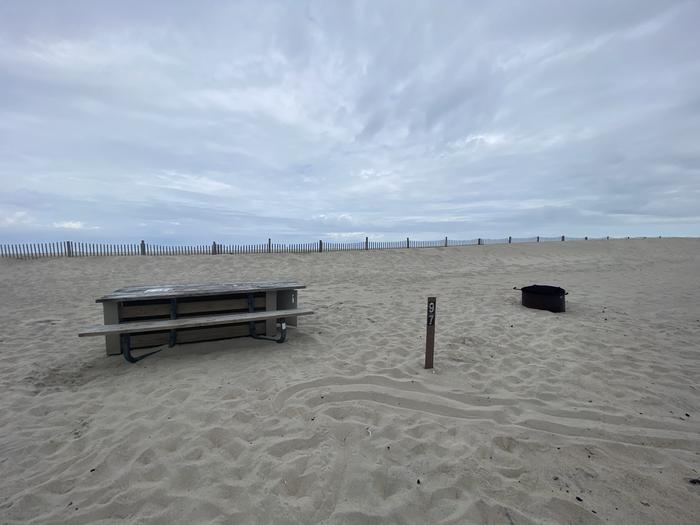Oceanside site 97 in May.  View of the wooden picnic table and black metal fire ring on the sand.  Sign post nearby that says 97 on it.  Dune fencing runs along the beach front on the horizon.Oceanside site 97 in May.