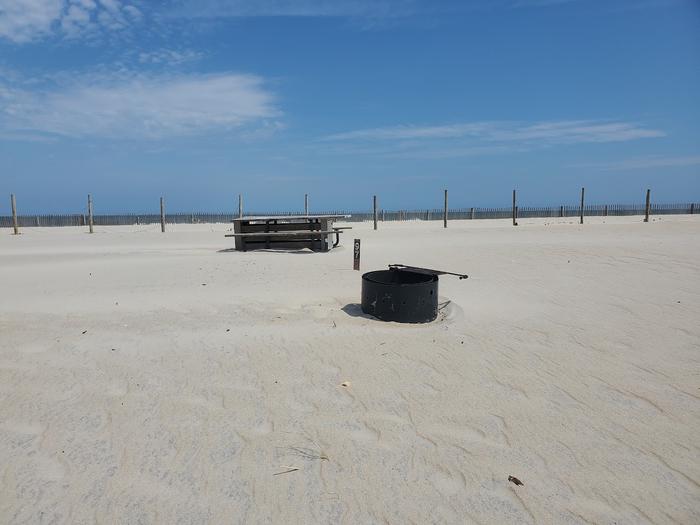 Oceanside site 97 in August.  View of the black metal fire ring on the sand.  Sign post and wooden picnic table are behind it.  Dune fencing runs along the beach front on the horizon.Oceanside site 97 in August.