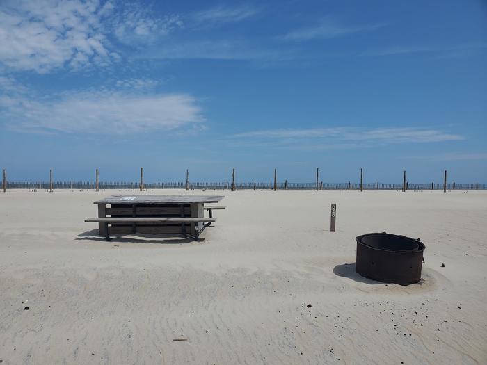 Oceanside site 98 in August.  View of black metal fire ring and wooden picnic table on the sand.  Sign post nearby says 98 on it.  Oceanside site 98 in August.