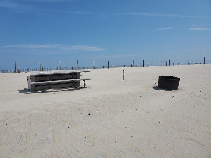 Oceanside site 98 in August.  View of the black metal fire ring and the wooden picnic table on the sand.  Sign post nearby says 98 on it.  Dune fencing runs along the beach front behind the campsite.Oceanside site 98 in August.