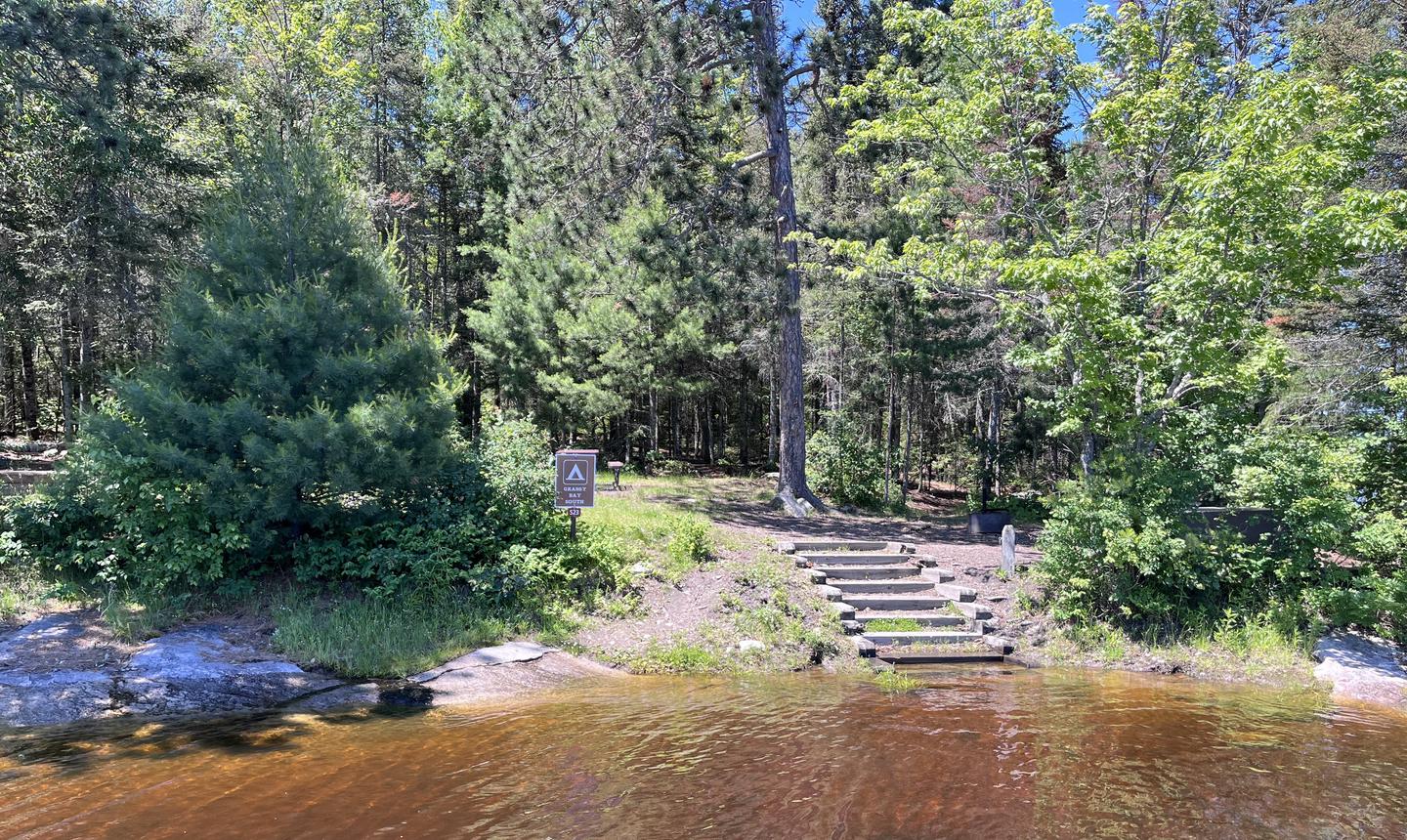 S23 - Grassy Bay South, view of boat access from water with stairs leading down to the shoreline and the campsite sign and mooring post on shoreView of campsite from the water