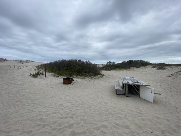 Oceanside site 100 in May.  View of the wooden picnic table and black metal fire ring on the sand.  Sign post nearby says 100 on it.  Dunes on the horizon.Oceanside site 100 in May.