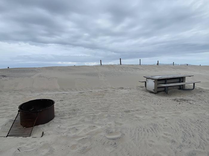 Oceanside site 103 in May.  View of the black metal fire ring and wooden picnic table on the sand.  Dune fencing runs along the beach front behind the campsite.  Sand has been built up behind the campsite.Oceanside site 103 in May.