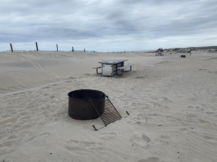 Oceanside site 103 in May.  Close view of the black metal fire ring on the sand.  Wooden picnic table is further into the campsite behind the fire ring.  Dune fencing runs along the beach front to the left.  Sand has been built up to the left of the campsite.  Other campsite within the view.Oceanside site 103 in May.