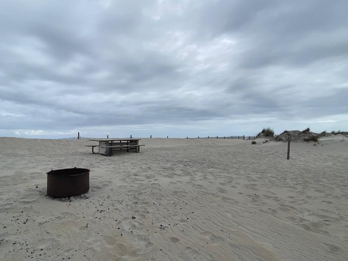 Oceanside site 104 in May.  View of the black metal fire ring on the sand.  Wooden picnic table is behind the fire ring further into the campsite.  Sign post nearby says 104 on it.  Dune fencing runs along the beach front on the horizon.Oceanside site 104 in May.