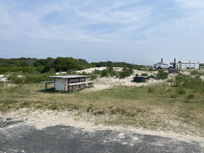Oceanside site 4 in July.  View of the wooden picnic table and black metal fire ring.  Bathrooms on the horizon to the left.  Brush surrounds the campsite.Oceanside site 4 in July.