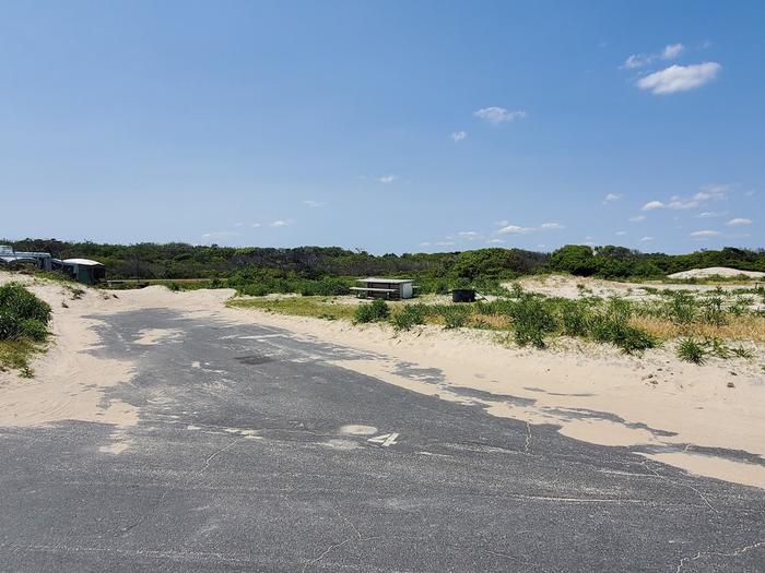 Oceanside site 4 in June.  View from the main road of the paved back in drive in site.  Wooden picnic table and black metal fire ring to the right of the pavement.  There is a spray painted 4 on the pavement at the entrance of the campsite.Oceanside site 4 in June.