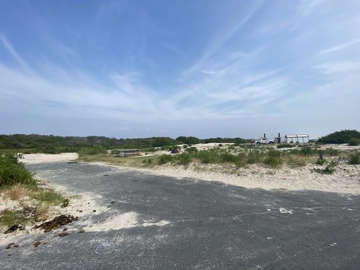 Oceanside site 4 in July.  View of the paved back in drive in site from the main road.  There is a white spray painted 4 on the pavement.  Wooden picnic table and black metal fire ring at the campsite.  Bathrooms on the horizon.Oceanside site 4 in July.