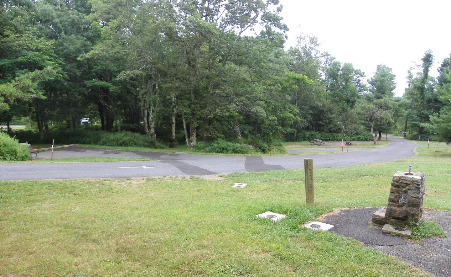 A stone water fountain next to a road that is lined with campsites. Each campsite is surrounded by trees and bushes.