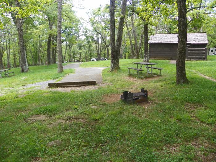 Site i220 grass lawn, with a short driveway, picnic table, and fire ring. In the background, a wood cabin building for restrooms. 