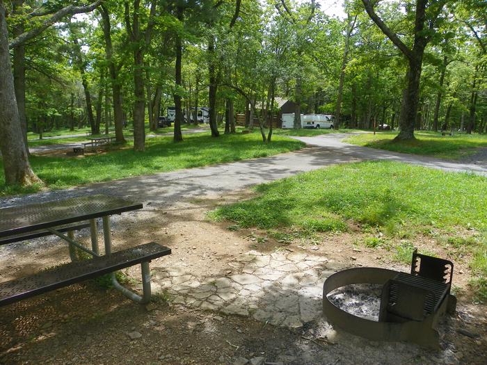 Site i225 picnic table and fire ring on a gravel pad. The pull through driveway exits onto the loop road, with a tree directly across from the exit. Restroom building and more campsites in the background.