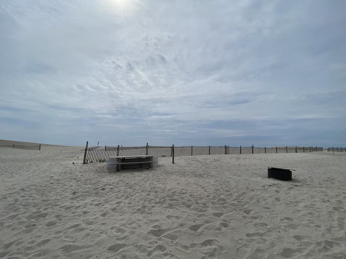 Oceanside site 51 in July 2024.  View of the wooden picnic table and black metal fire ring on the sand.  Dune fencing runs along the beach front behind the campsite.  There is an opening leading to a pathway.Oceanside site 51 - July 2024.