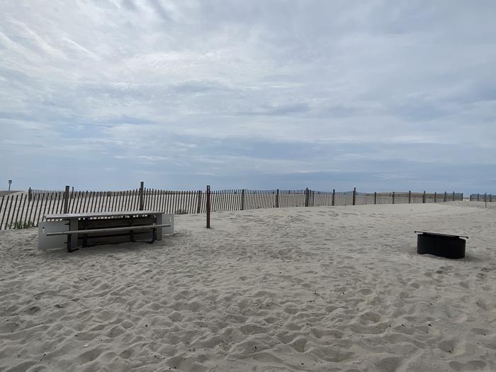 Oceanside site 51 in July 2024.  View of the wooden picnic table and black metal fire ring on the sand.  Dune fencing runs along the beach front behind the campsite.  Oceanside site 51 - July 2024.