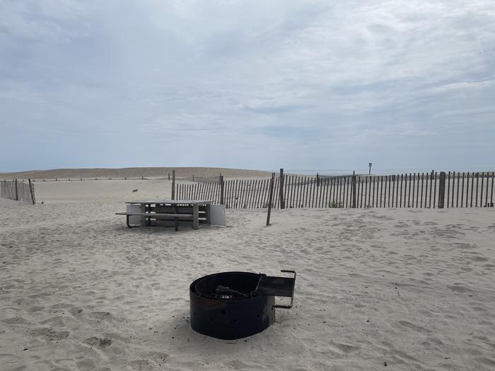 Oceanside site 51 in July 2024.  View of the black metal fire ring on the sand.  Wooden picnic table and signpost nearby behind the fire ring.  Dune fencing runs along the beach front behind the campsite.  There is an opening nearby that leads to a walkway that leads out to the ocean front.Oceanside site 51 - July 2024. 