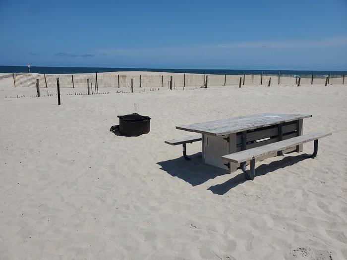 Oceanside site 51 in July 2023.  View of the wooden picnic table and black metal fire ring on the sand.  Dune fencing runs along the beach front behind the campsite.  Ocean front on the horizon.  Oceanside site 51 - July 2023.