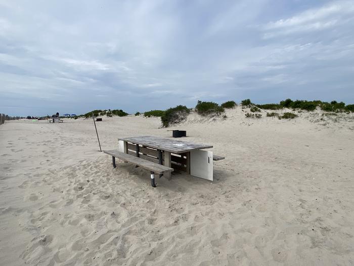 Oceanside site 51 in July 2024.  View of the wooden picnic table on the sand.  Black metal fire ring is further behind the campsite.  Signpost nearby says "51" on it.Oceanside site 51 - July 2024.