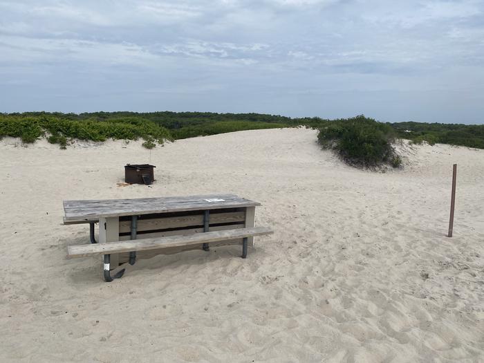 Oceanside site 52 in July 2024.  View of the wooden picnic table on the sand.  Signpost nearby says "52" on it.  Black metal fire ring is further behind the picnic table.Oceanside site 52 - July 2024.