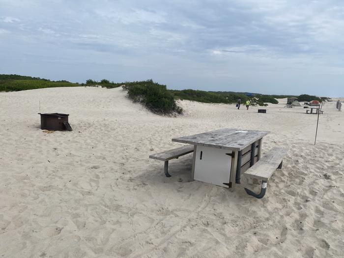 Oceanside site 52 in July 2024.  View of the wooden picnic table and black metal fire ring on the sand.  Signpost nearby in the sand.  Brush behind the campsite on the horizon.  Other campers and campsites within the view.Oceanside site 52 - July 2024.