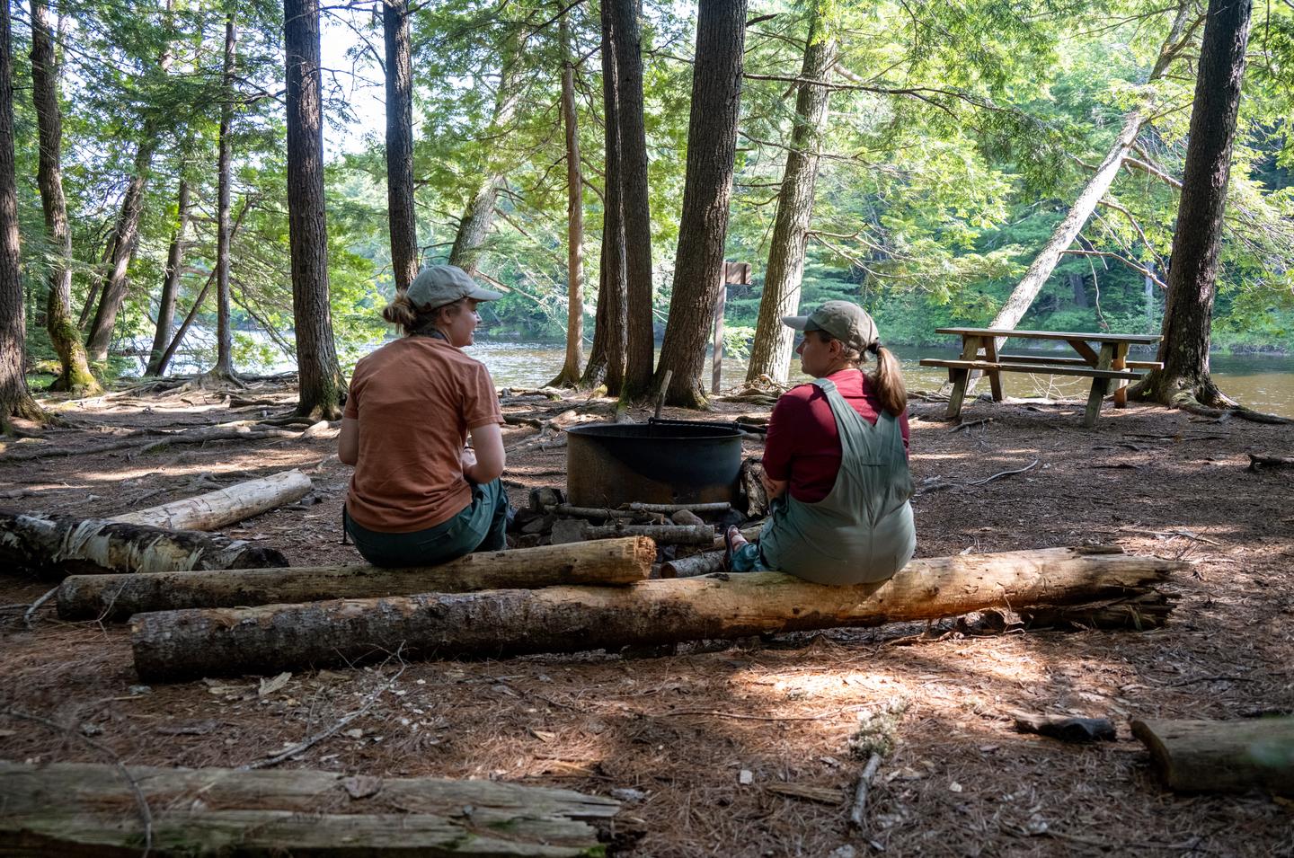 Two women campers sit in a shaded campsite on fallen logs around a black metal fire ring with a view of the river. A wooden picnic table is just to the right ahead of them.Enjoy the scenic view of the East Branch of the Penobscot River at Big Spring Brook Campsite.