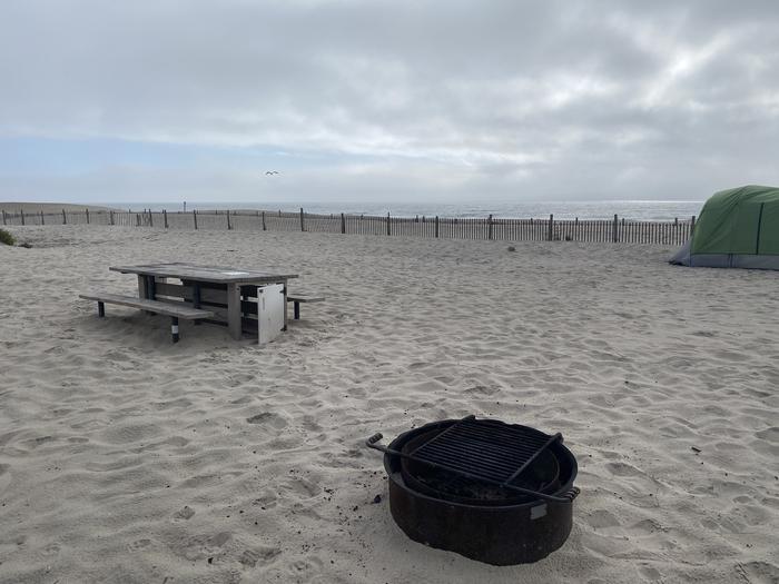 Oceanside site 55 in August 2024.  View of the black metal fire ring and wooden picnic table on the sand.  Dune fencing runs along the beach front behind the campsite.  Another camper's tent is within the view.  Ocean front on the horizon.Oceanside site 55 - August 2024.