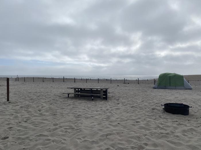 Oceanside site 55 in August 2024.  View of the wooden picnic table and black metal fire ring on the sand.  Signpost nearby says "55" on it.  Other camper's tent within the view.  Dune fencing runs along the beach behind the campsite with ocean front on the horizon.Oceanside site 55 - August 2024.