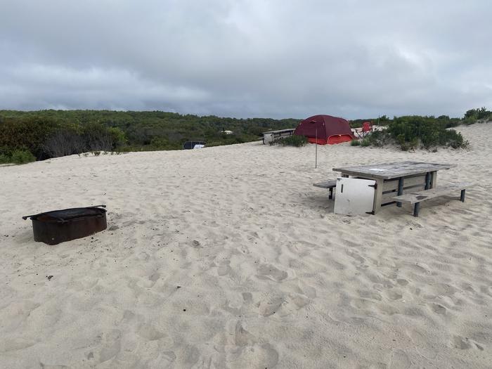 Oceanside site 55 in August 2024.  View of the wooden picnic table and black metal fire ring on the sand.  Other camper's tents within the view.  Brush on the horizon.Oceanside site 55 - August 2024.