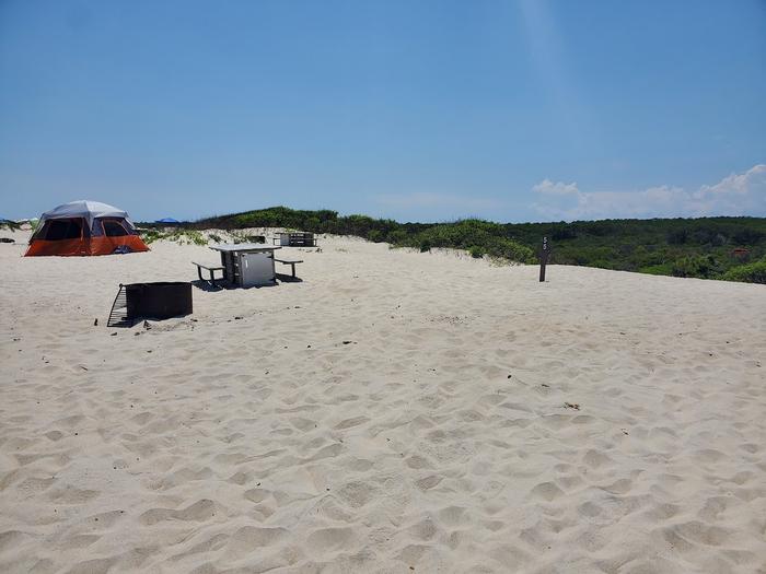 Oceanside site 55 in July 2023.  View of the black metal fire ring and wooden picnic table on the sand.  Signpost nearby says "55" on it.  Other camper's tents within the view.  Brush on the horizon.Oceanside site 55 - July 2023.