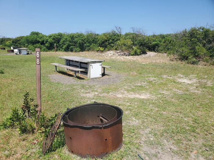 Oceanside site 68 in July 2023.  Close up view of the black metal fire ring on the grass.  Signpost nearby says "68" on it.  Wooden picnic table is behind the fire ring.  Other campsites within the view.  Brush on the horizon. Oceanside site 68 - July 2023.