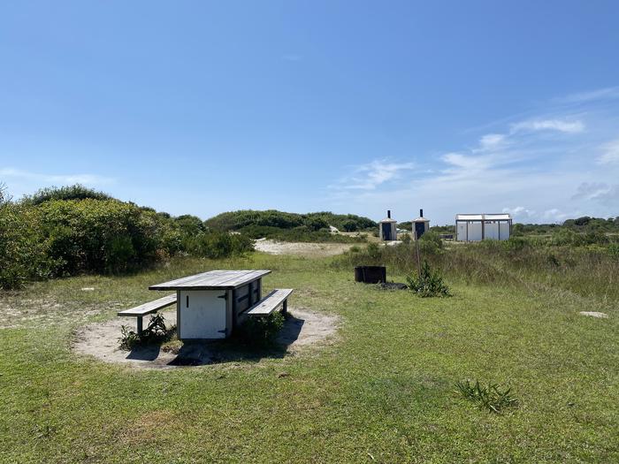 Oceanside site 68 in August 2024.  View of the wooden picnic table and black metal fire ring on the grass.  Bathrooms are within the view on the horizon.  Brush surrounds the campsite.Oceanside site 68 - August 2024. 