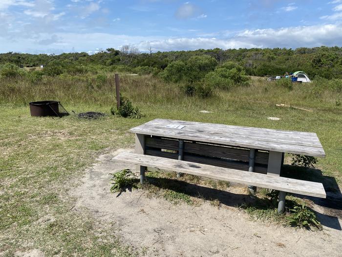 Oceanside site 68 in August 2024.  View of the wooden picnic table and black metal fire ring on the grass.  Signpost nearby says "68" on it.  Other campsites and campers' tents within the view.  Brush surrounds the campsite on the horizon.Oceanside site 68 - August 2024.