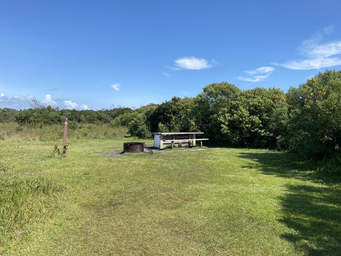 Oceanside site 70 in August 2024.  View of the wooden picnic table and black metal fire ring on the grass.  Signpost nearby says "70" on it.  Brush surrounds the campsite.Oceanside site 70 - August 2024.