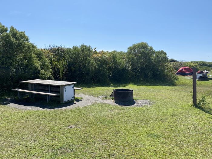 Oceanside site 70 in August 2024.  View of the black metal fire ring and wooden picnic table on the grass.  Signpost nearby.  Other camper's tents within the view.  Brush behind the campsite.Oceanside site 70 - August 2024.