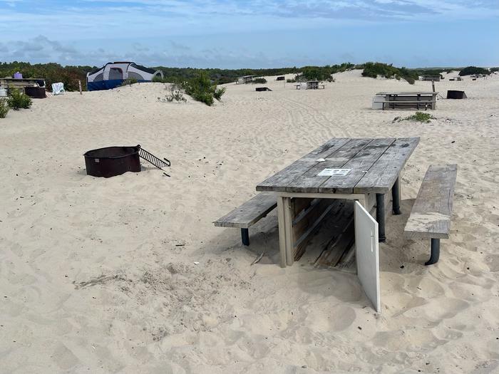 Oceanside site 58 in September 2024.  View of the wooden picnic table and black metal fire ring on the sand.  Other campsites and campers equipment within the view.Oceanside site 58 - September 2024.