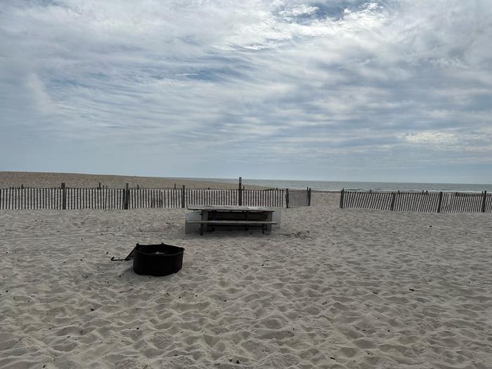 Oceanside site 58 in September 2024.  View of the black metal fire ring and wooden picnic table on the sand.  There is dune fencing along the beach front behind the campsite.  There is an opening leading to the ocean front in the fencing. Oceanside site 58 - September 2024.