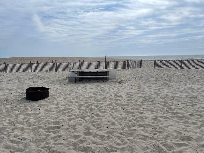 Oceanside site 58 in September 2024. View of the wooden picnic table and black metal fire ring on the sand.  There is dune fencing running along the beach front behind the campsite.  There is a break in the fencing that leads to the ocean front.  Ocean on the horizon.Oceanside site 58 - September 2024.