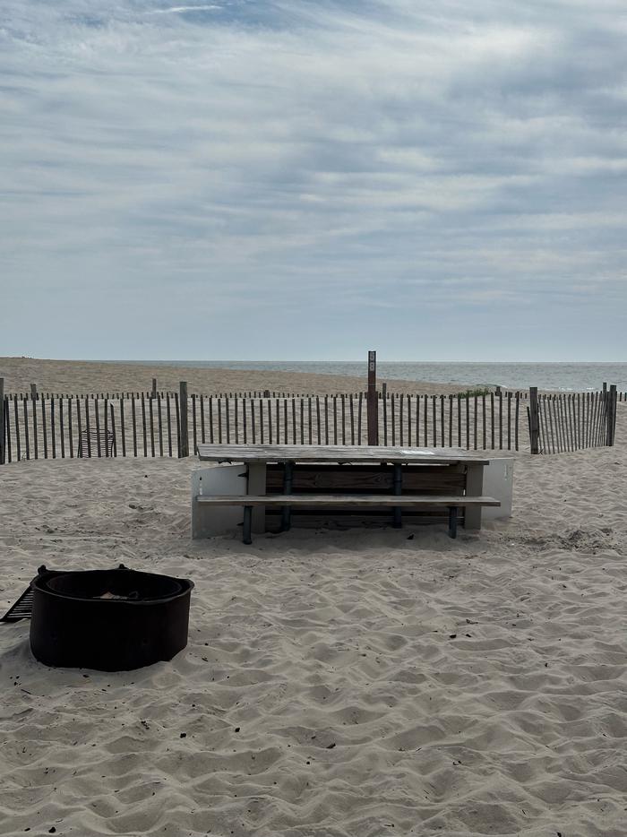 Oceanside site 58 in September 2024.  View of the wooden picnic table and black metal fire ring on the sand.  Dune fencing runs along the beach front behind the campsite.  Signpost nearby says "58" on it.  Ocean front within the view.Oceanside site 58 - September 2024.