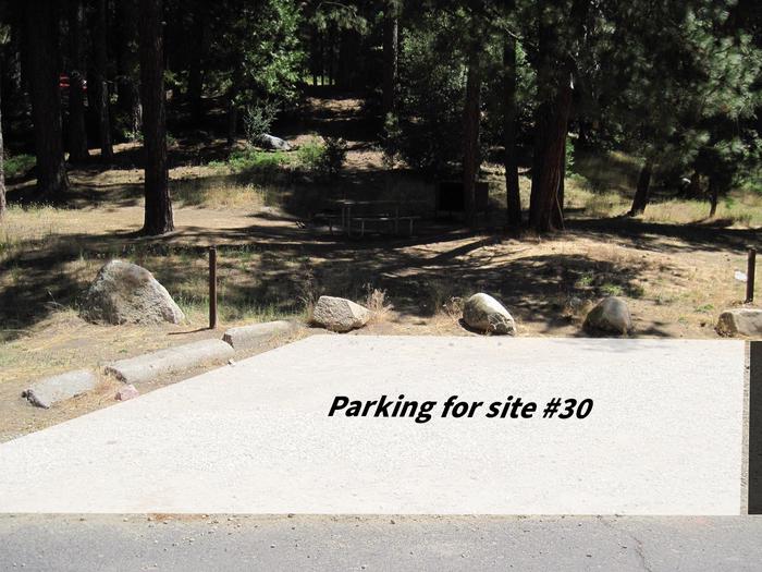 A small section of gravel parking pad at the bottom of grassy hill. It is bordered by rocks and logs. Overlayed on the image is a white box, indicating the parking for site 30Parking is shared for sites #28, #29, and #30. There is space for 2 cars to park here.