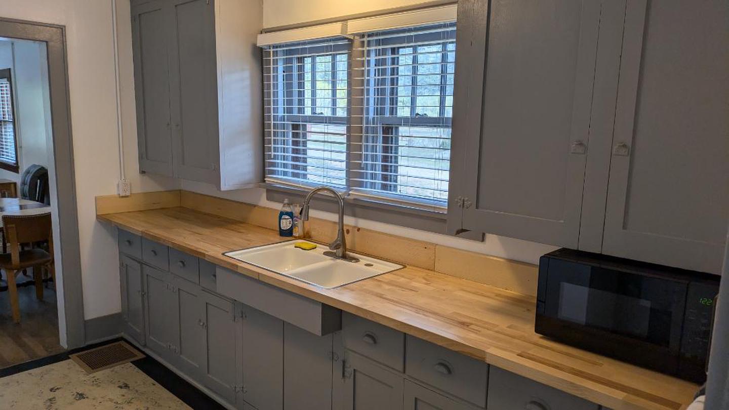 A photo of kitchen cabinets and countertop with an enamel double sink and black microwave. 