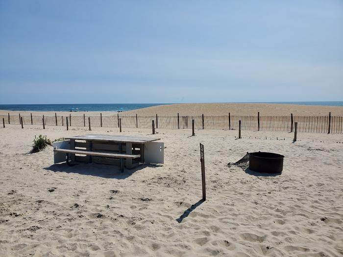 Oceanside site 58 in August 2023.  View of the wooden picnic table and black metal fire ring on the sand.  Signpost between them says "58" on it.  Dune fencing runs along the beach front behind the campsite.  Partial view of the ocean front on the horizon. Oceanside site 58 - August 2023.