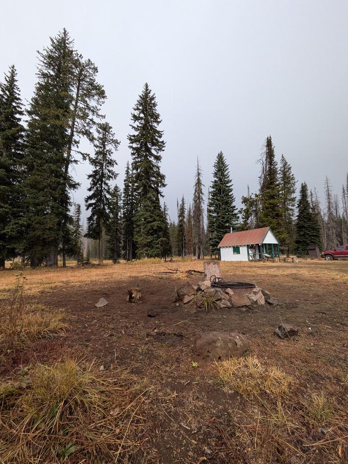 Fire ring with Buck Park Cabin and tall pine trees in the background. Fire pit and Buck Park Cabin