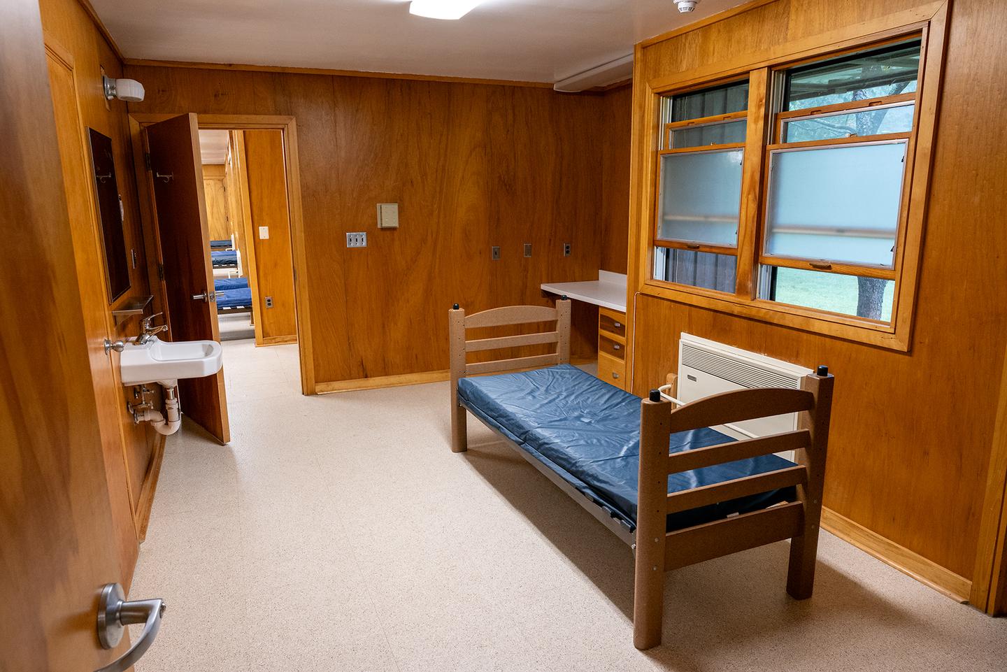 Individual dormitory room, vinyl floor, wooden walls, and one double window. One single bed with vinyl covered mattress, metal storage unit, and small porcelain wall sink.                                                                                There are 2 individual rooms in each of the 4 dormitories.  These rooms contain a single bed with vinyl mattress, a closet, metal storage unit, small sink, wall mirror and wall mounted writing area (not visible)